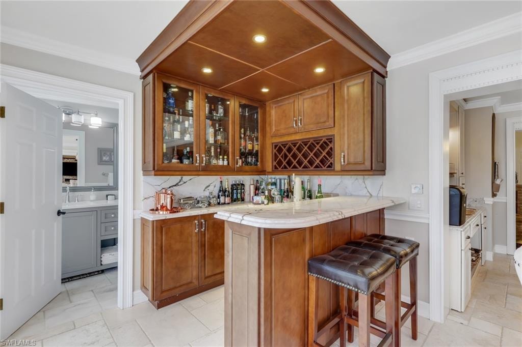 Indoor bar with ornamental molding, brown cabinetry, tasteful backsplash, glass insert cabinets, and recessed lighting