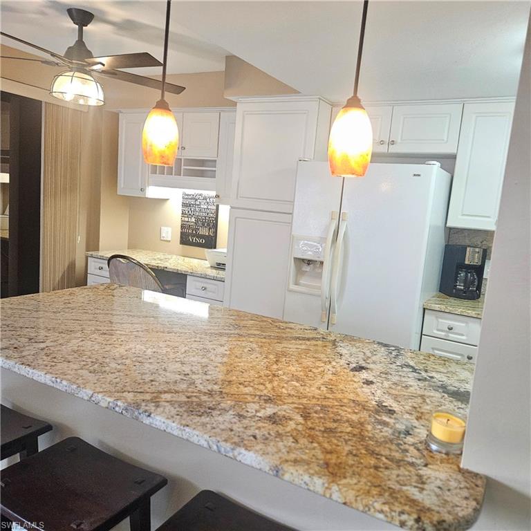 Kitchen with white cabinetry, a kitchen breakfast bar, light stone countertops, white fridge with ice dispenser, and hanging light fixtures