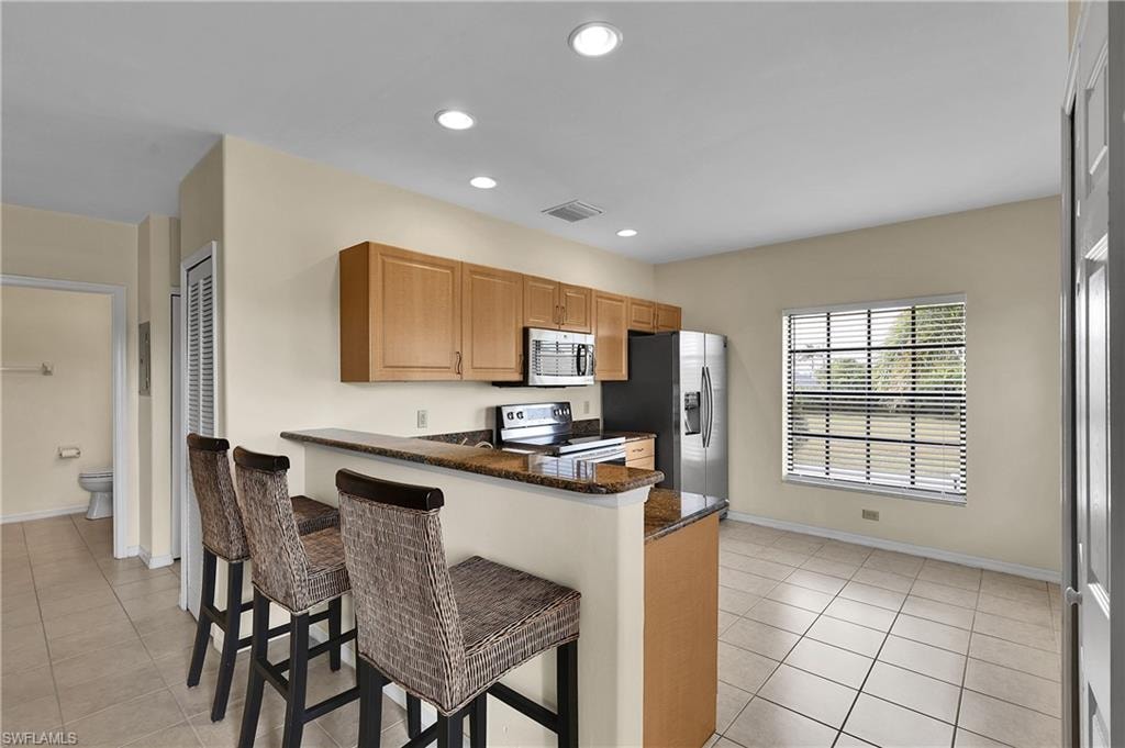 Kitchen with light tile patterned floors, appliances with stainless steel finishes, dark stone countertops, recessed lighting, and a breakfast bar