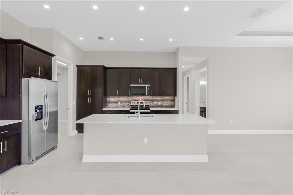 Kitchen featuring stainless steel appliances, dark brown cabinets, decorative backsplash, a kitchen island with sink, and recessed lighting