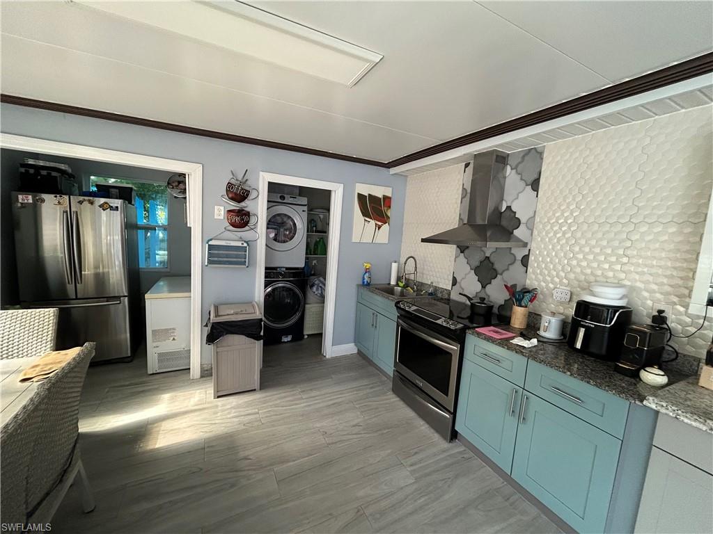 Kitchen featuring appliances with stainless steel finishes, wall chimney exhaust hood, stacked washer and clothes dryer, tasteful backsplash, and dark stone counters