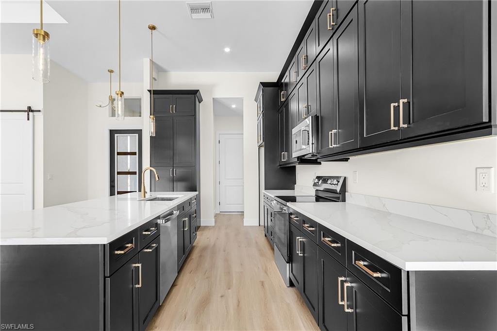 Kitchen with dark cabinets, stainless steel appliances, light stone counters, an island with sink, and pendant lighting