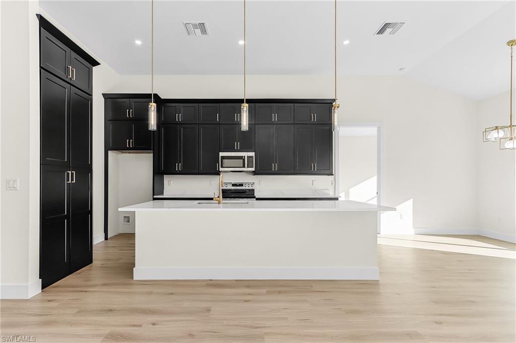 Kitchen with dark cabinets, light wood-style floors, stainless steel appliances, an island with sink, and vaulted ceiling
