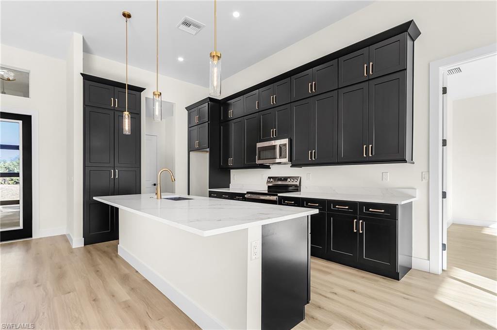 Kitchen featuring dark cabinetry, a center island with sink, hanging light fixtures, and light stone counters