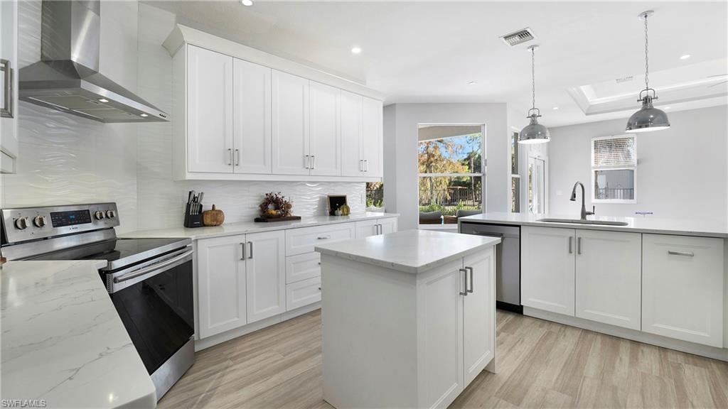 Kitchen featuring stainless steel appliances, wall chimney exhaust hood, a kitchen island, decorative light fixtures, and white cabinetry