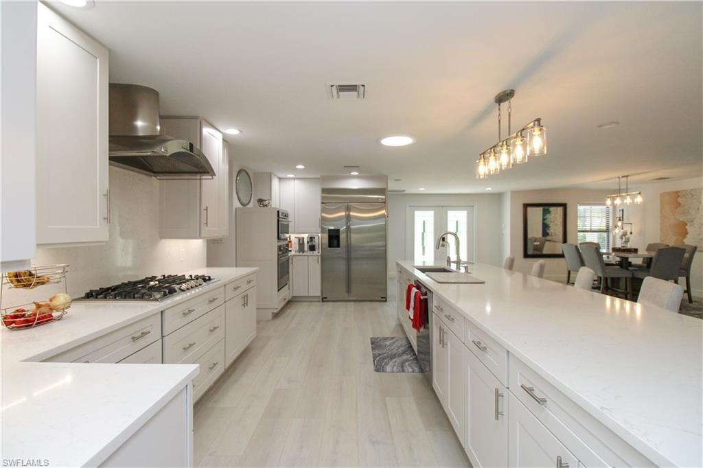 Kitchen featuring wall chimney exhaust hood, hanging light fixtures, appliances with stainless steel finishes, white cabinets, and light wood-type flooring