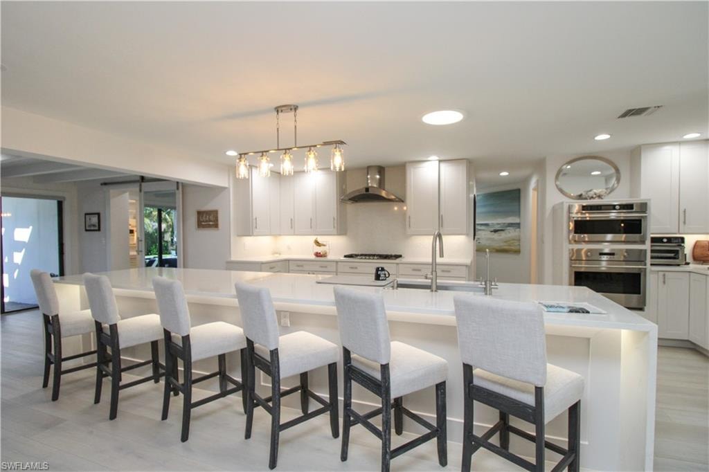 Kitchen featuring pendant lighting, a breakfast bar, a center island with sink, wall chimney range hood, and double oven