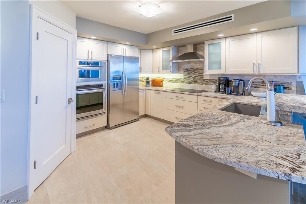 Kitchen with light stone countertops, appliances with stainless steel finishes, glass insert cabinets, white cabinets, and wall chimney exhaust hood