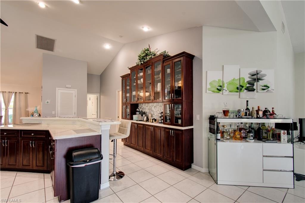 Indoor dry bar featuring dark brown cabinetry, glass insert cabinets, vaulted ceiling, light tile patterned floors, and light stone countertops