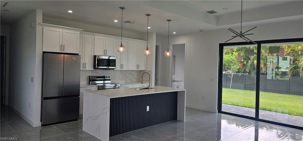 Kitchen with appliances with stainless steel finishes, white cabinetry, light stone countertops, decorative light fixtures, and a center island with sink