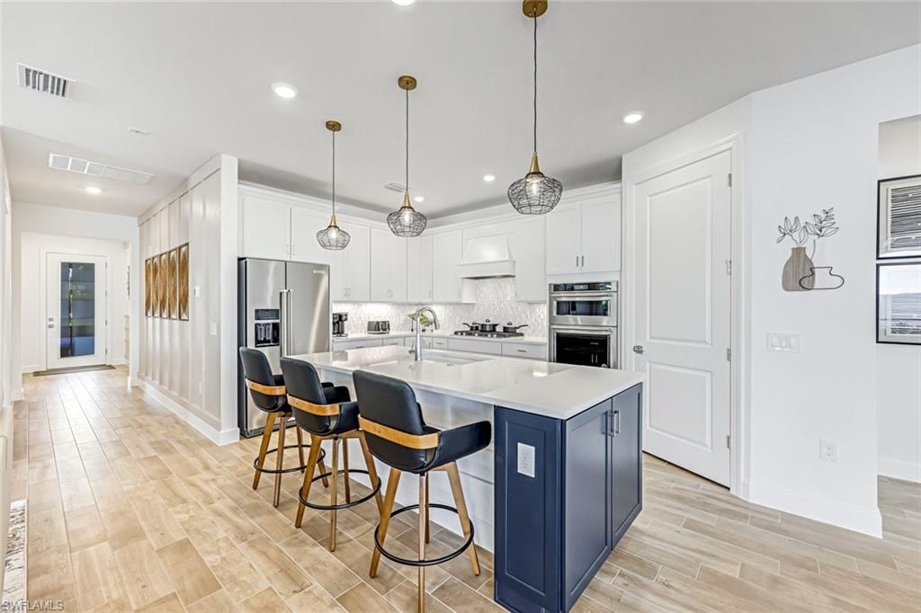 Kitchen featuring white cabinets, stainless steel appliances, a kitchen island with sink, wood tiled floors, and pendant lighting