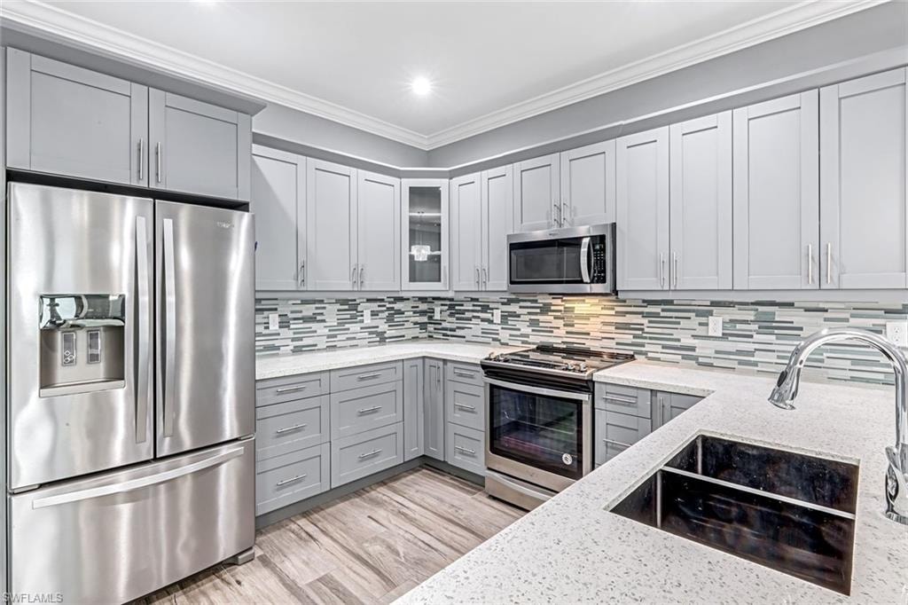 Kitchen with stainless steel appliances, gray cabinets, ornamental molding, glass fronted cabinets, and light stone counters
