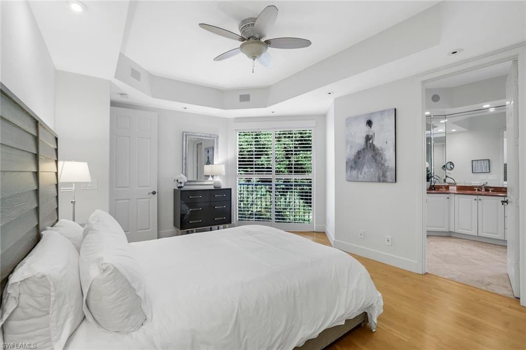 Bedroom with light wood-style flooring, ceiling fan, ensuite bathroom, recessed lighting, and a raised ceiling