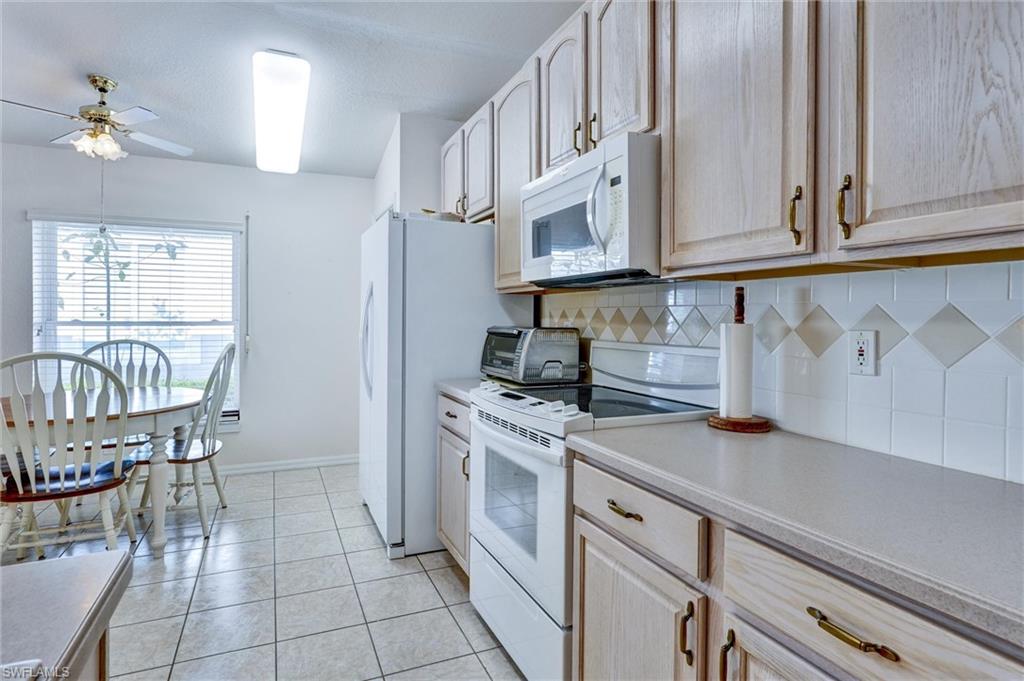 Kitchen featuring light wood-finish cabinetry with brass-tone hardware, white appliances, tiled backsplash with decorative accents, and light-toned countertops