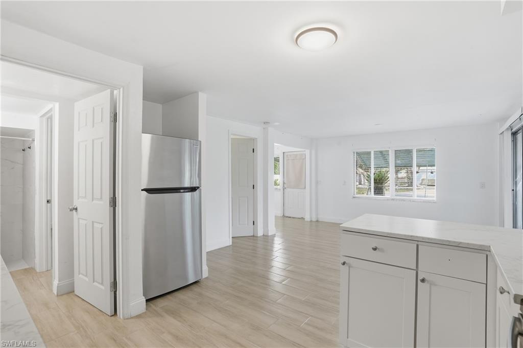 Open-concept living area featuring light wood-finish flooring, white cabinetry with a light countertop, a stainless steel refrigerator, and multiple windows - Virtually Edited Image