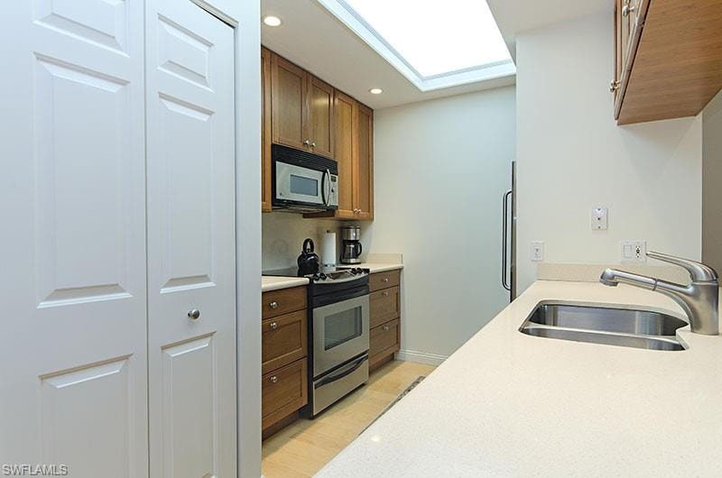 Kitchen with electric stove, white microwave, recessed lighting, brown cabinetry, and light stone countertops