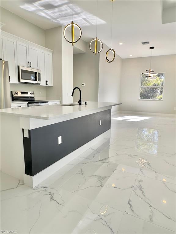 Kitchen featuring hanging light fixtures, appliances with stainless steel finishes, white cabinetry, light marble finish flooring, and a kitchen island with sink