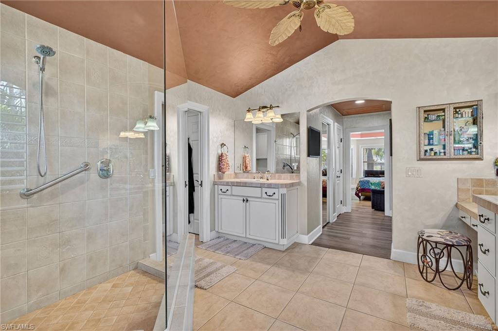 Ensuite bathroom featuring vanity, light tile patterned floors, a walk in shower, vaulted ceiling, and ceiling fan