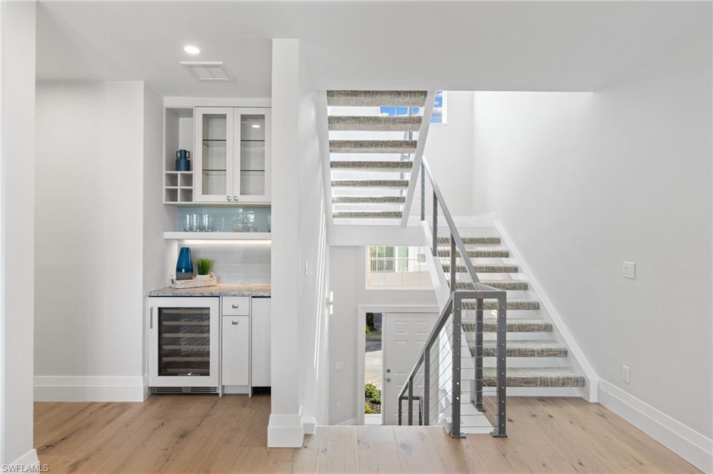 Indoor dry bar with white cabinets, beverage cooler, glass insert cabinets, light wood finished floors, and stairway