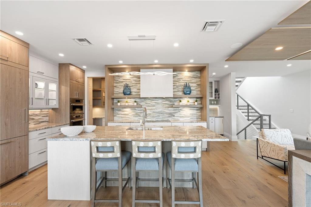 Kitchen featuring light stone counters, pendant lighting, open shelves, a center island with sink, and a breakfast bar area