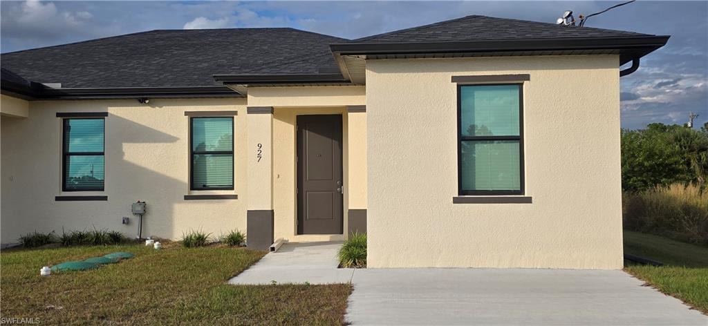 View of front facade with roof with shingles, a front yard, and stucco siding