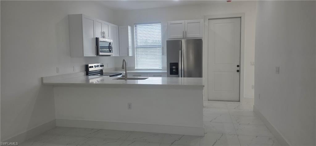 Kitchen featuring appliances with stainless steel finishes, white cabinetry, a peninsula, light stone countertops, and light marble finish flooring