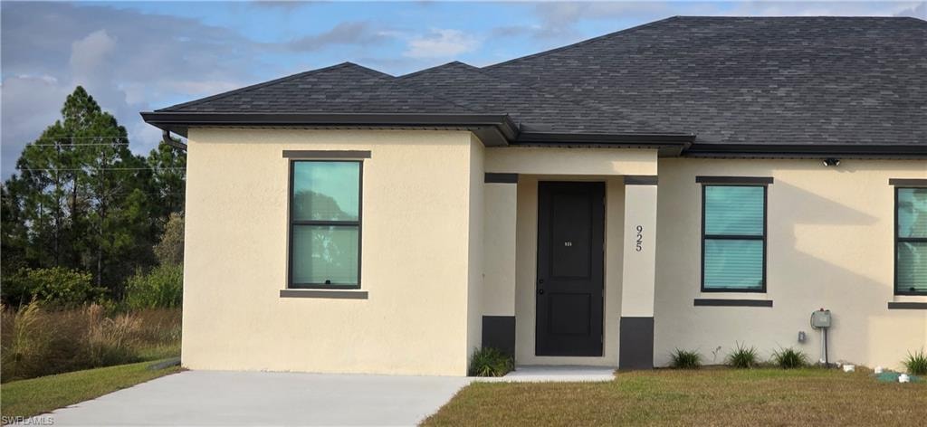 View of exterior entry with roof with shingles, stucco siding, and a lawn