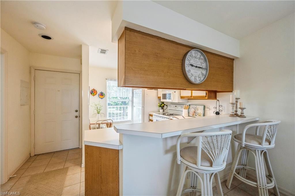 Kitchen with light countertops, a peninsula, a breakfast bar, white appliances, and brown cabinetry