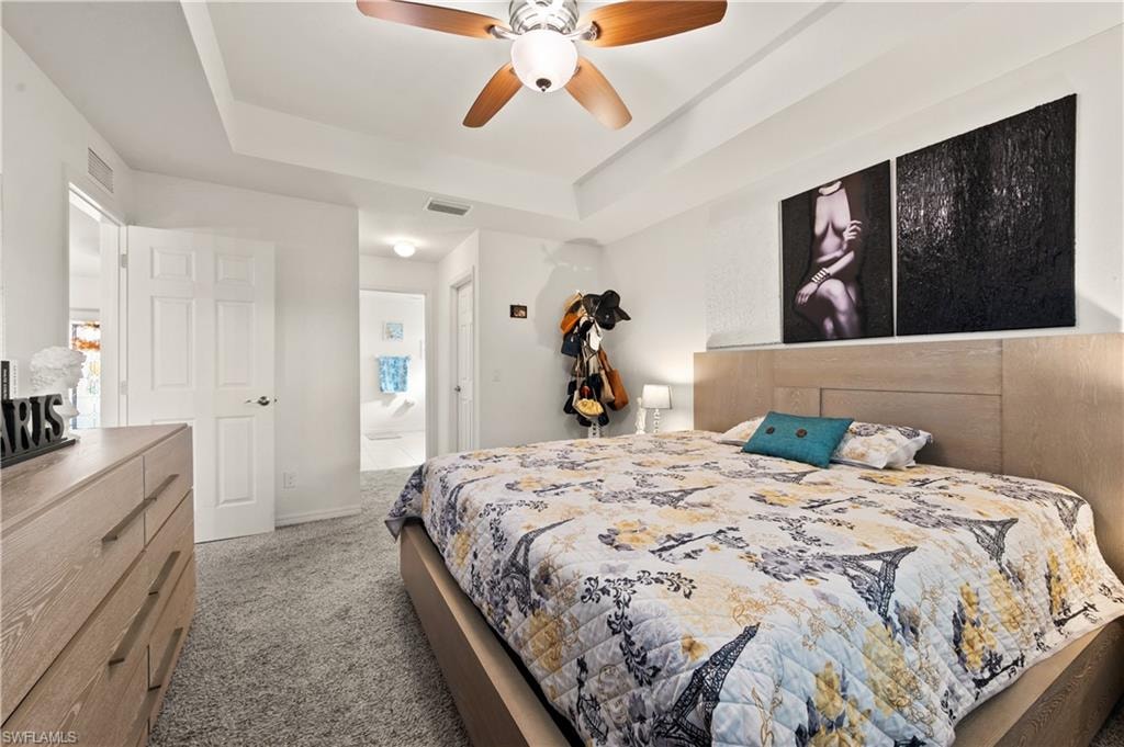 Bedroom with light colored carpet, a raised ceiling, a ceiling fan, and connected bathroom