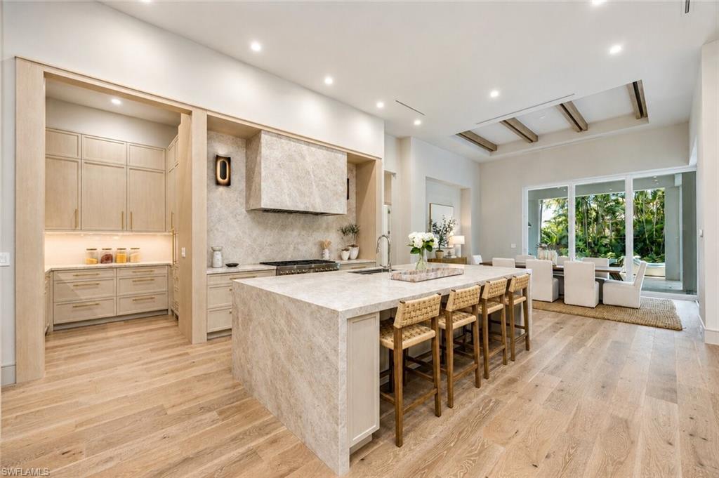 Open-concept kitchen featuring an expansive island with waterfall edge, wood-finish flooring, integrated range hood, recessed lighting, and a separate pantry area