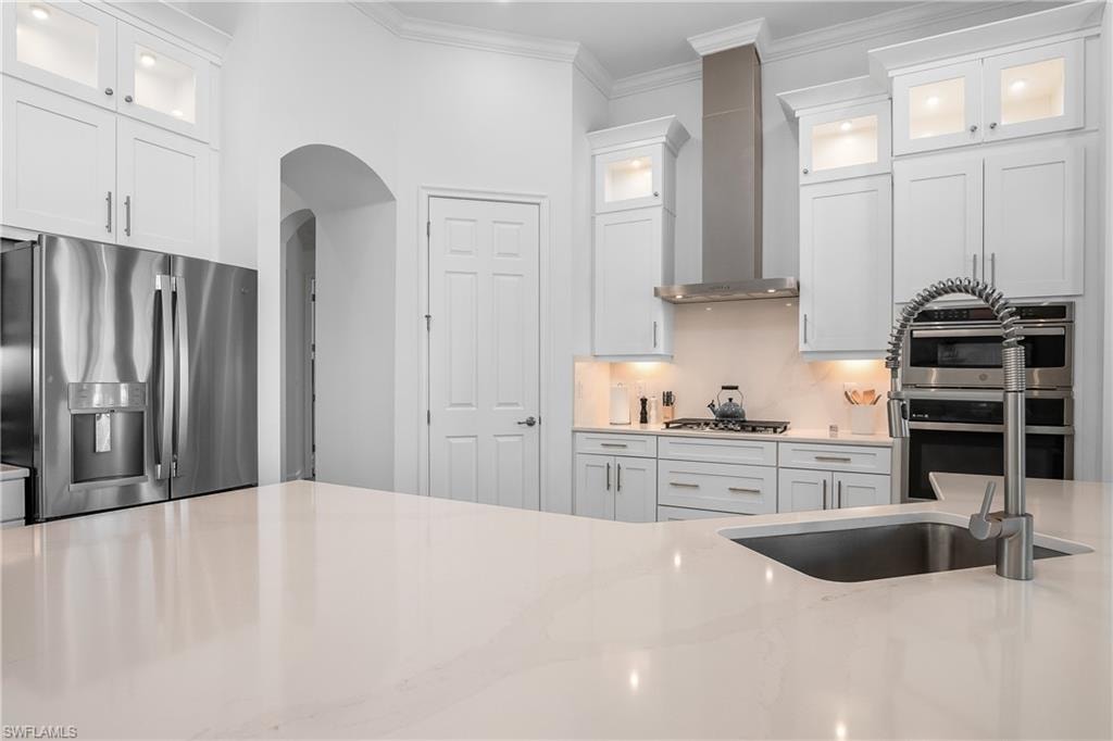 Kitchen featuring stainless steel appliances, glass insert cabinets, white cabinetry, ornamental molding, and wall chimney range hood