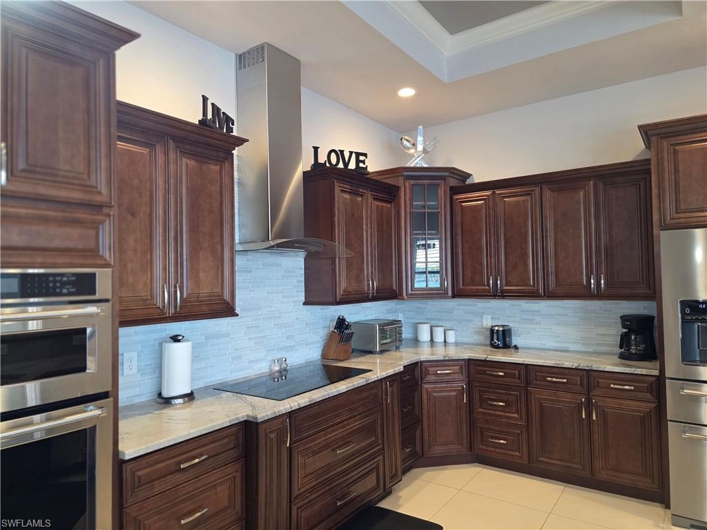 Kitchen featuring stainless steel appliances, dark brown cabinets, exhaust hood, a tray ceiling, and light stone counters