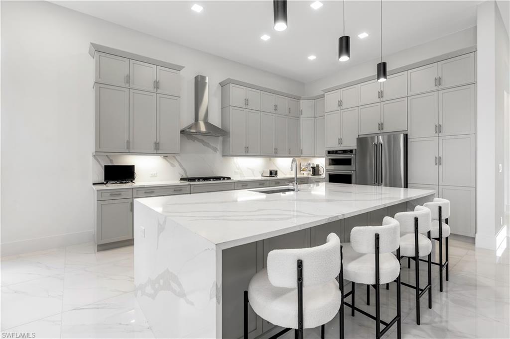 Kitchen featuring gray cabinetry, light stone counters, wall chimney exhaust hood, a kitchen bar, and stainless steel appliances