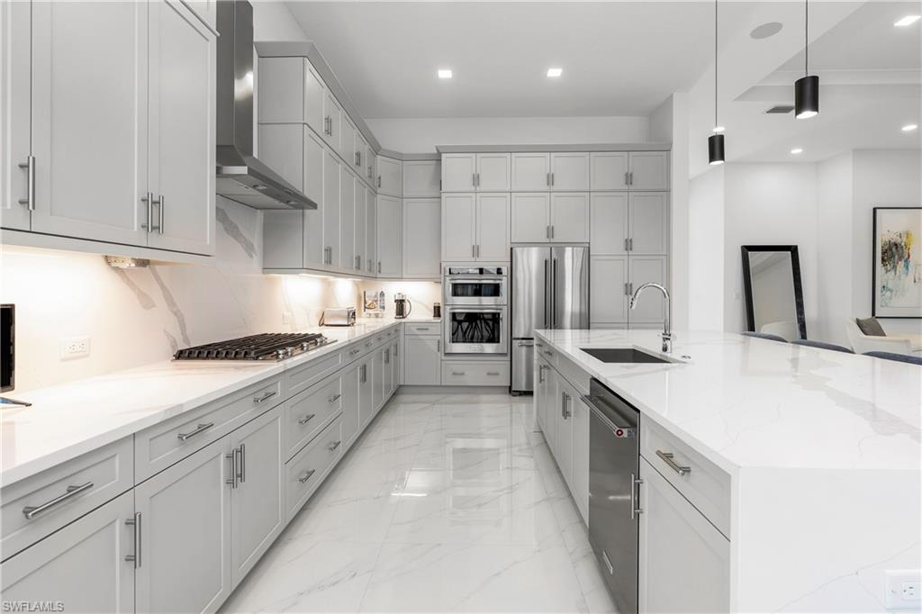 Kitchen featuring wall chimney range hood, decorative light fixtures, light stone counters, appliances with stainless steel finishes, and a large island with sink