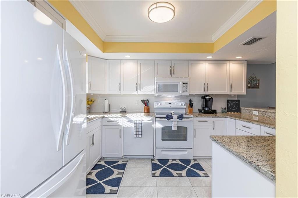 Kitchen with white appliances, ornamental molding, light stone counters, white cabinets, and tasteful backsplash