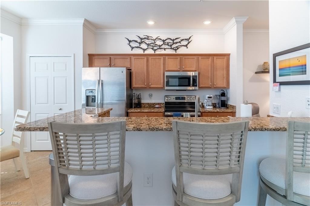 Kitchen with a kitchen bar, appliances with stainless steel finishes, crown molding, light stone countertops, and brown cabinets