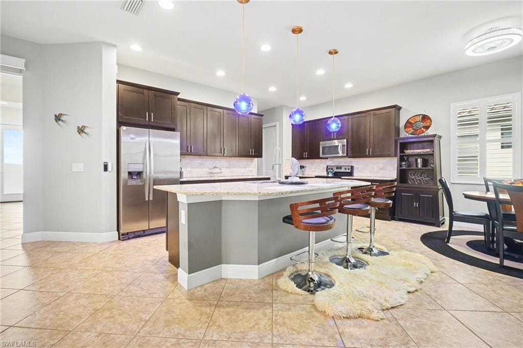 Kitchen featuring dark brown cabinets, appliances with stainless steel finishes, a breakfast bar area, light stone countertops, and decorative light fixtures