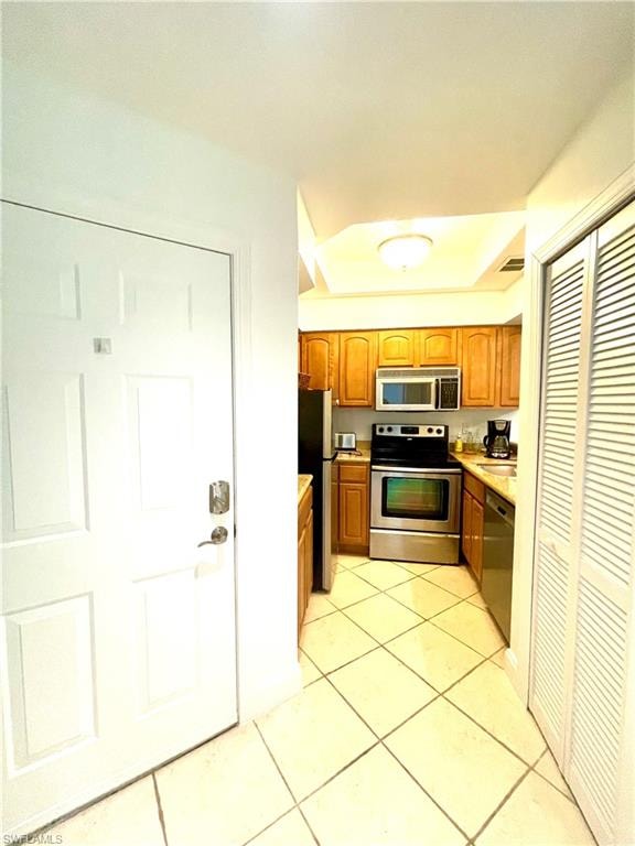 Kitchen featuring appliances with stainless steel finishes, light countertops, light tile patterned flooring, brown cabinetry, and a tray ceiling