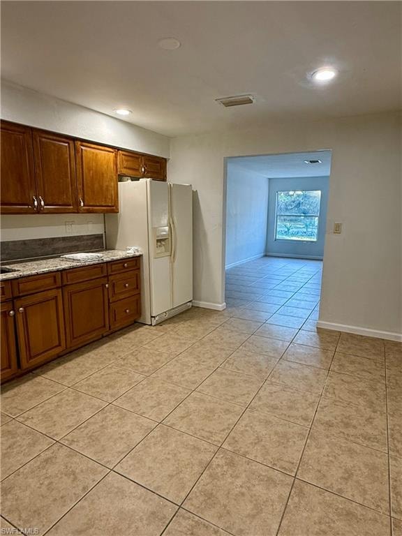 Kitchen with white refrigerator with ice dispenser, brown cabinetry, light tile patterned floors, light stone counters, and recessed lighting