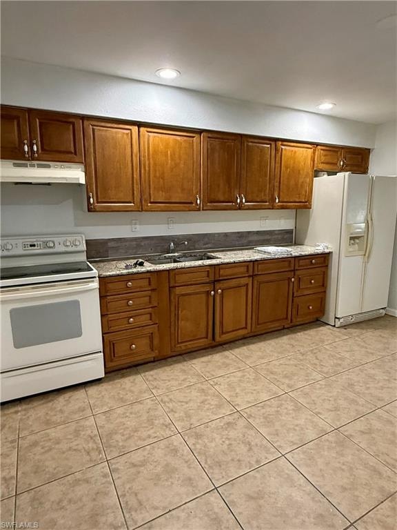 Kitchen featuring white appliances, brown cabinetry, under cabinet range hood, light tile patterned flooring, and recessed lighting