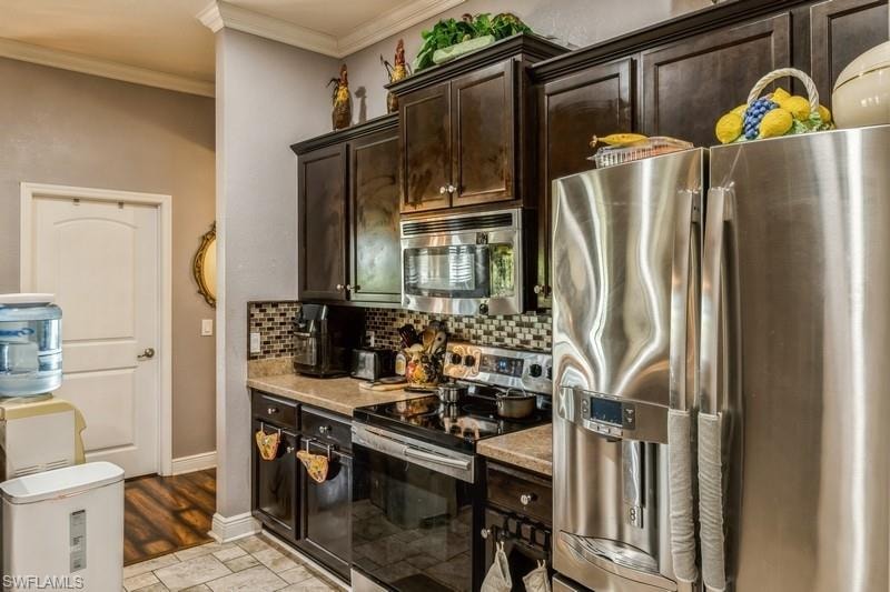 Kitchen featuring stainless steel appliances, dark brown cabinets, crown molding, decorative backsplash, and light stone countertops