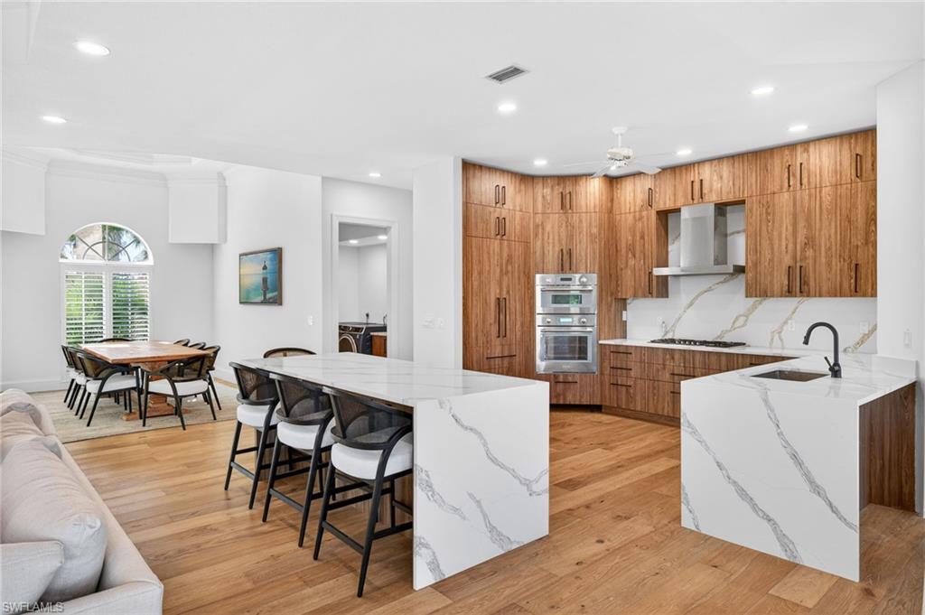 Kitchen featuring light stone counters, brown cabinets, a breakfast bar, open floor plan, and wall chimney exhaust hood