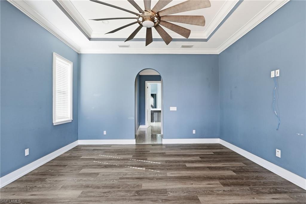 Empty room with a raised ceiling, arched walkways, dark wood-style floors, a ceiling fan, and ornamental molding
