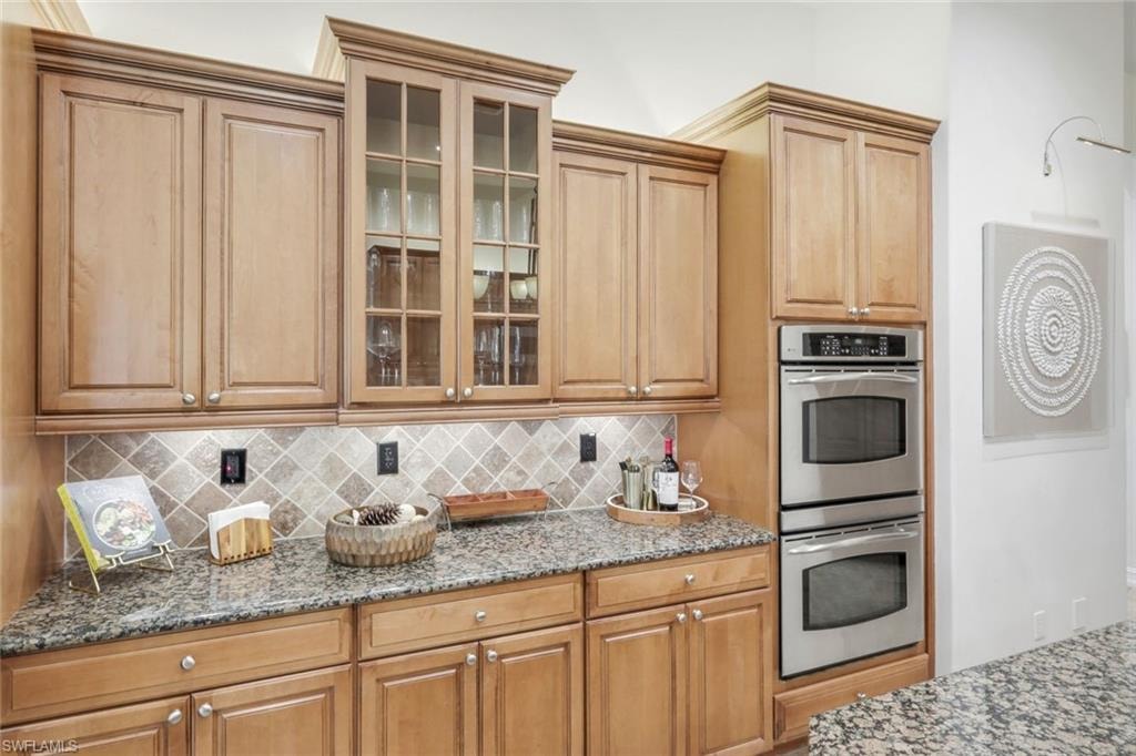 Kitchen with stainless steel double oven, glass fronted cabinets, dark stone counters, and backsplash