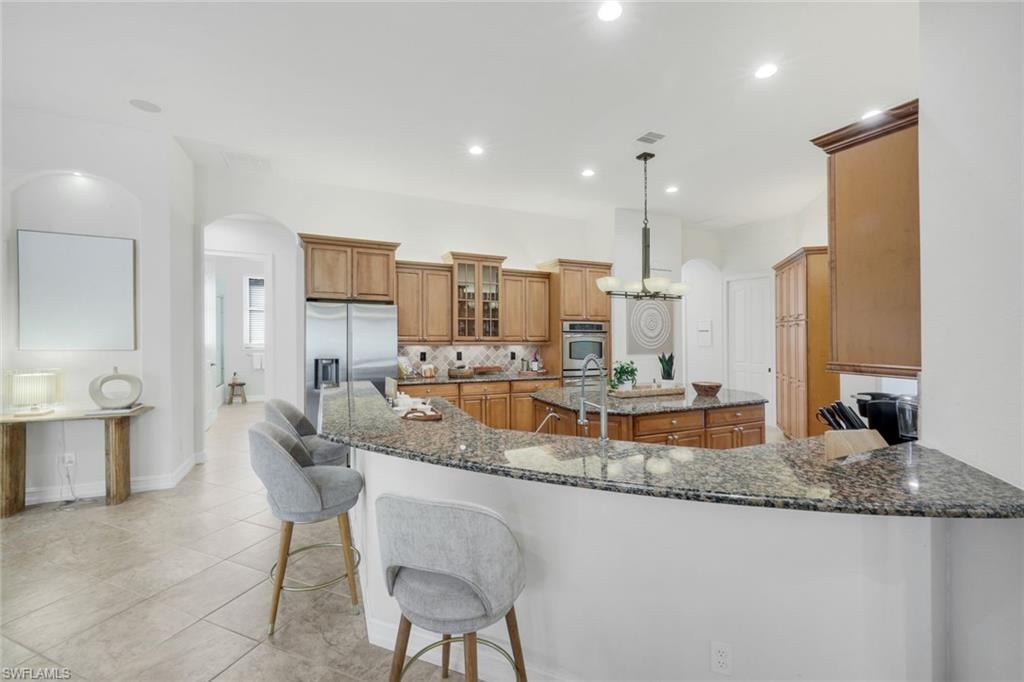 Kitchen with arched walkways, wood finish cabinets, dark stone counters, hanging light fixtures, and backsplash