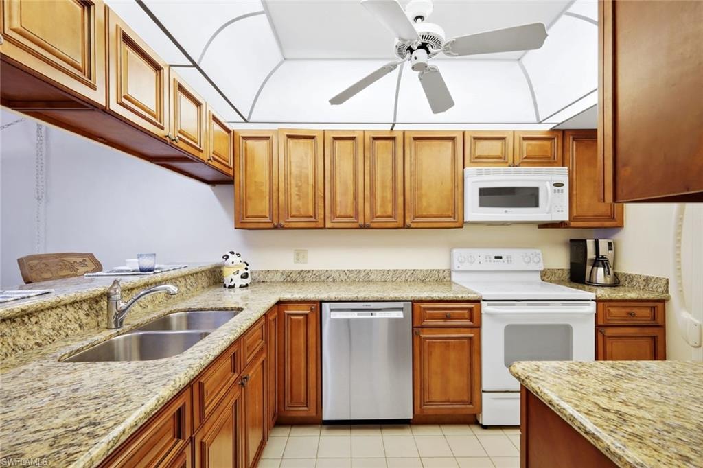 Kitchen with white appliances, light stone counters, ceiling fan, and brown cabinetry