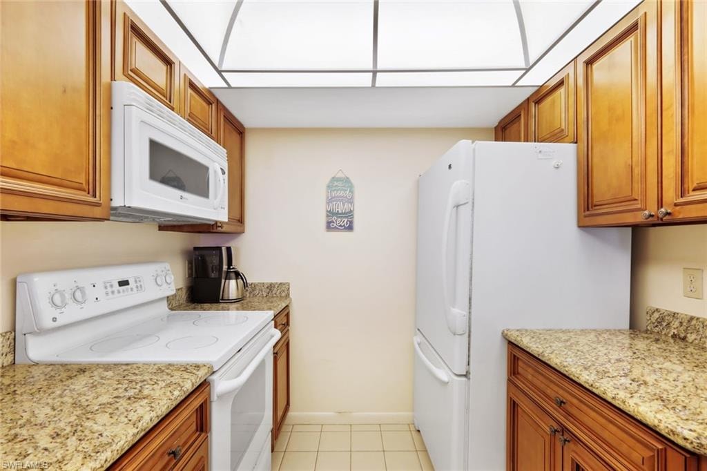 Kitchen featuring white appliances, brown cabinetry, light stone countertops, and light tile patterned floors