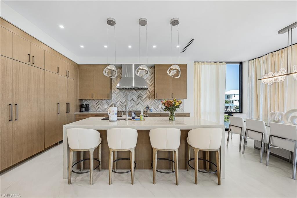 Kitchen with hanging light fixtures, a kitchen bar, light stone countertops, backsplash, and a center island with sink - Virtually Edited Image