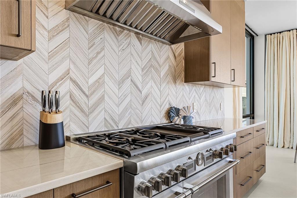 Kitchen featuring custom exhaust hood, range with two ovens, light stone countertops, modern cabinets, and tasteful backsplash - Virtually Edited Image
