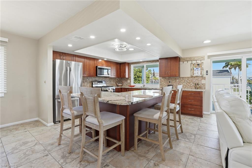 Kitchen with stainless steel appliances, light stone counters, brown cabinets, a kitchen island, and recessed lighting - Virtually Edited Image